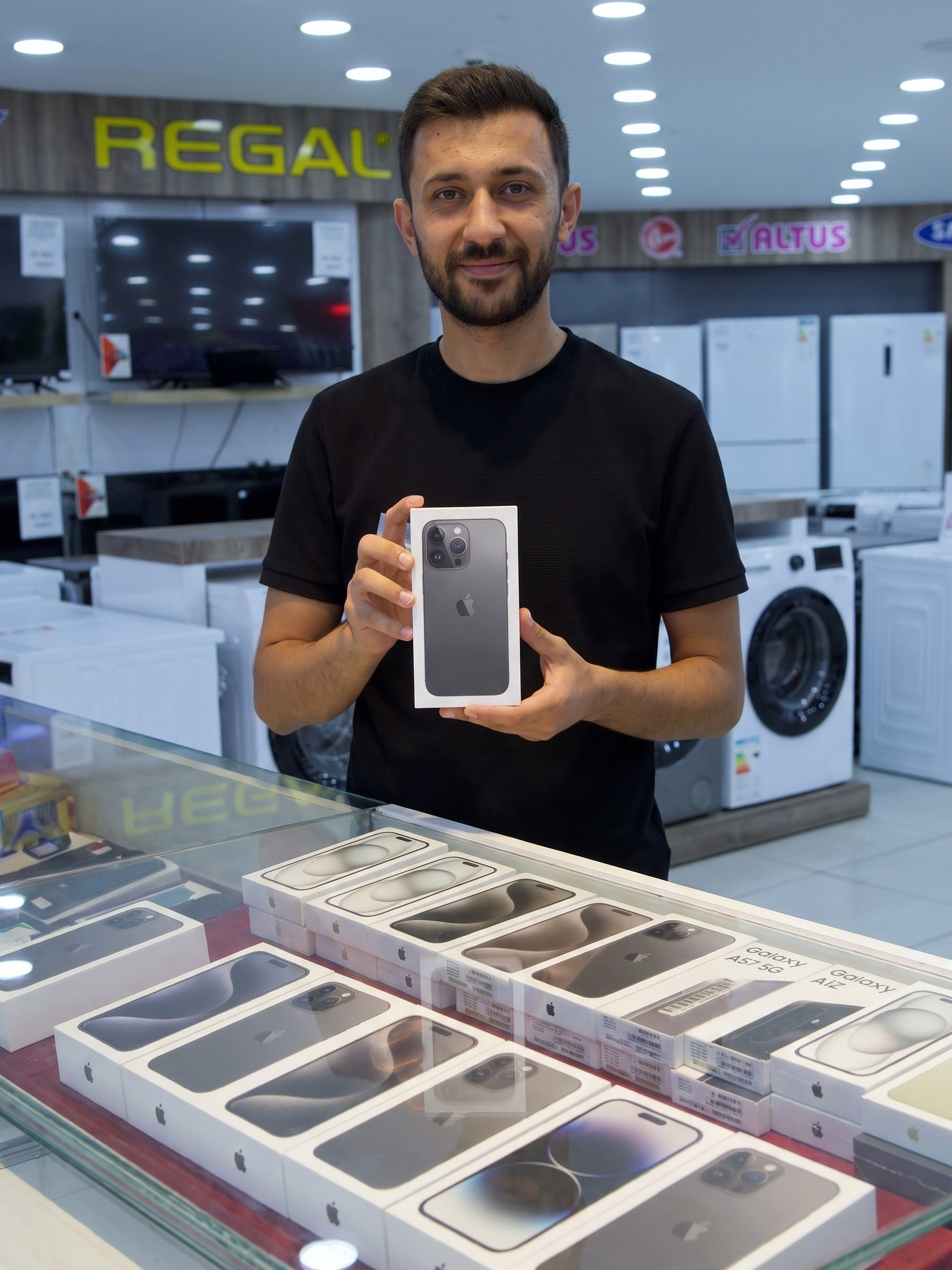 Mağaza çalışanı cam telefon bankosunun arkasında kutulu bir telefonu iki eliyle tutuyor, bankoda kutulu telefonlar dizili